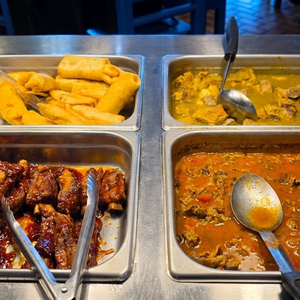A buffet spread with trays of BBQ ribs, plantains, chicken curry, and a rich red stew.