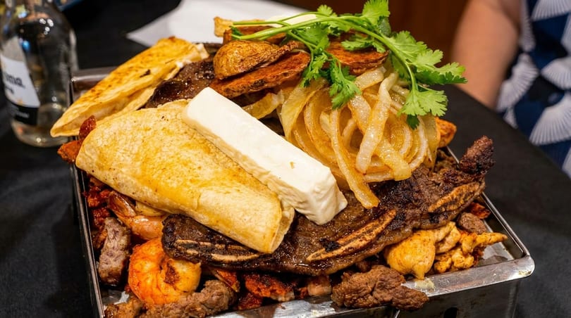 A large platter of grilled meats, shrimp, tortillas, cheese, onions, and potatoes, garnished with cilantro.