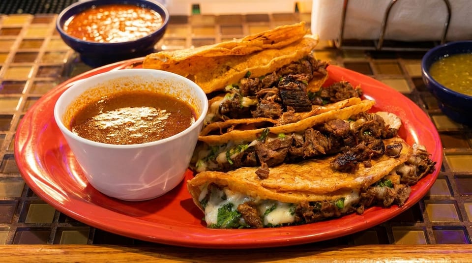 A red plate holds several cheesy birria tacos with a side of consommé and salsa.